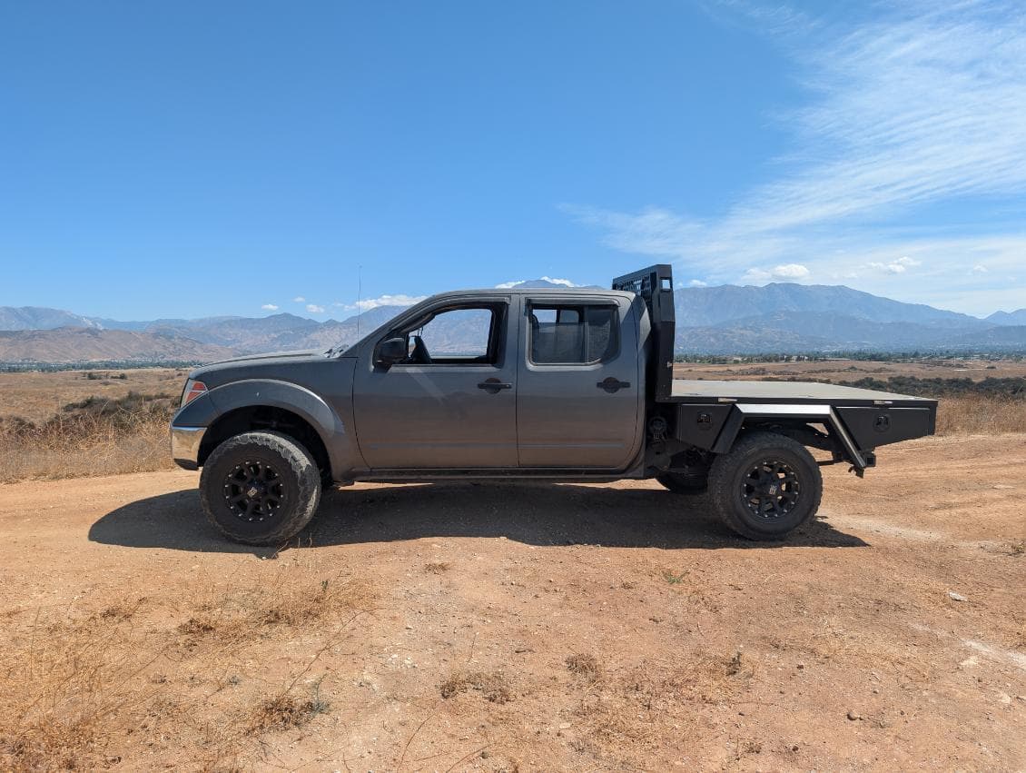 Grey Nissan Frontier with a black aluminum flatbed tray and mesh headboard, parked on a dirt ridge with mountains behind