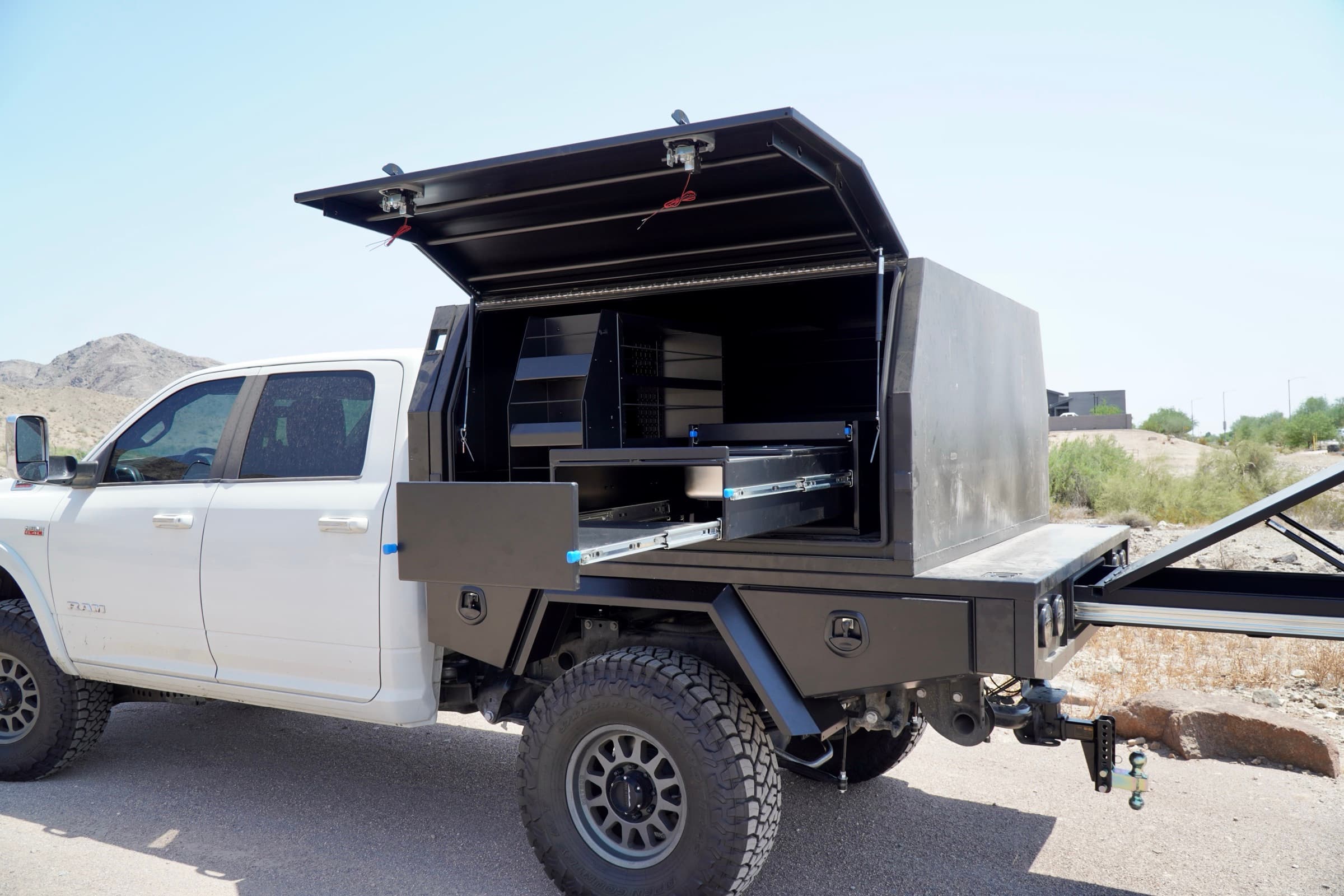 White Ram 3500 with a black aluminum flatbed tray and touring canopy, gullwing door raised showing internal shelving and an extended slide-out drawer