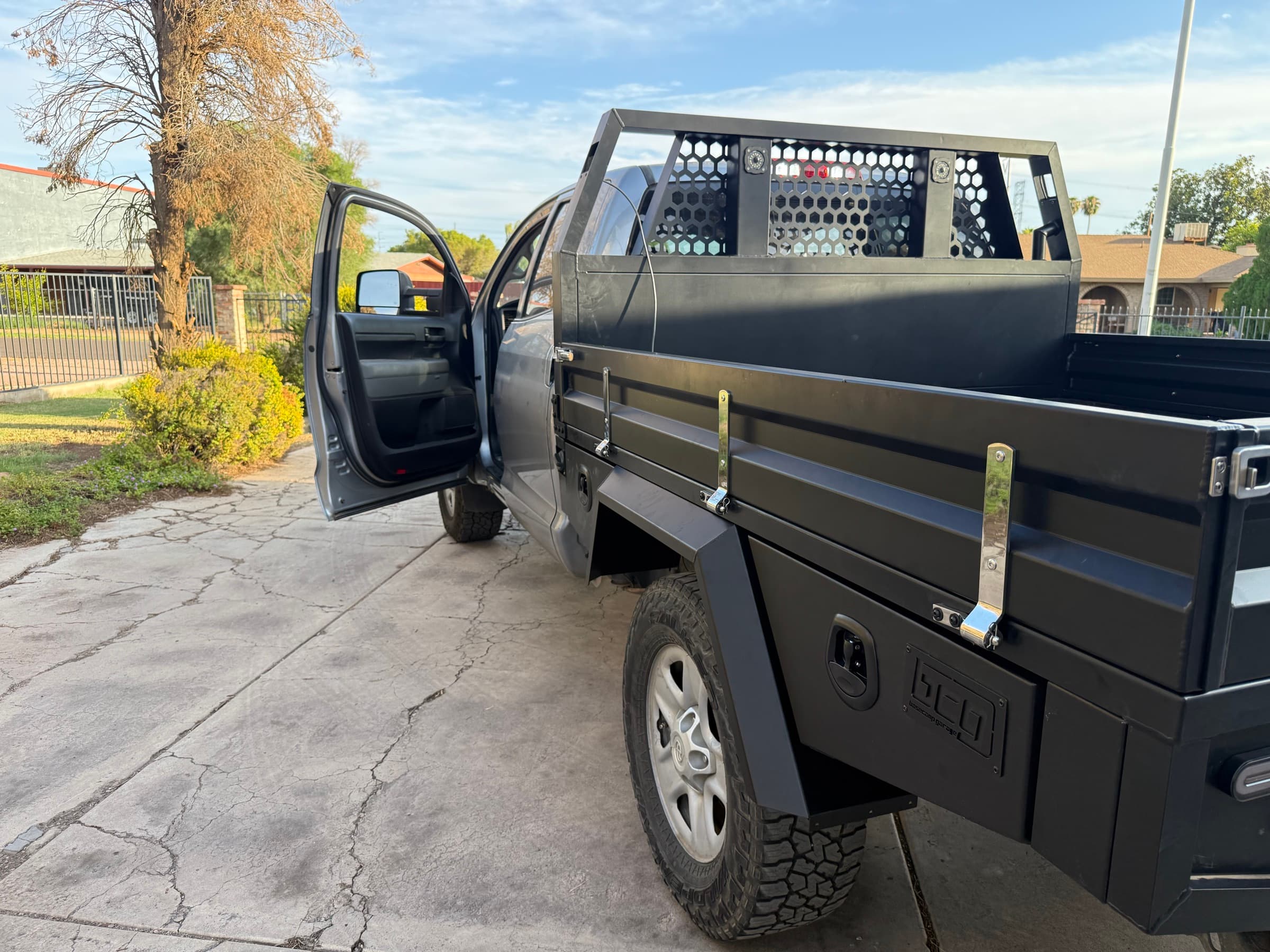 Closeup of the 2013 Tundra flatbed showing the hex-mesh headboard with integrated lights, polished stainless dropside latches, and a Basecamp Garage stamped under-tray toolbox