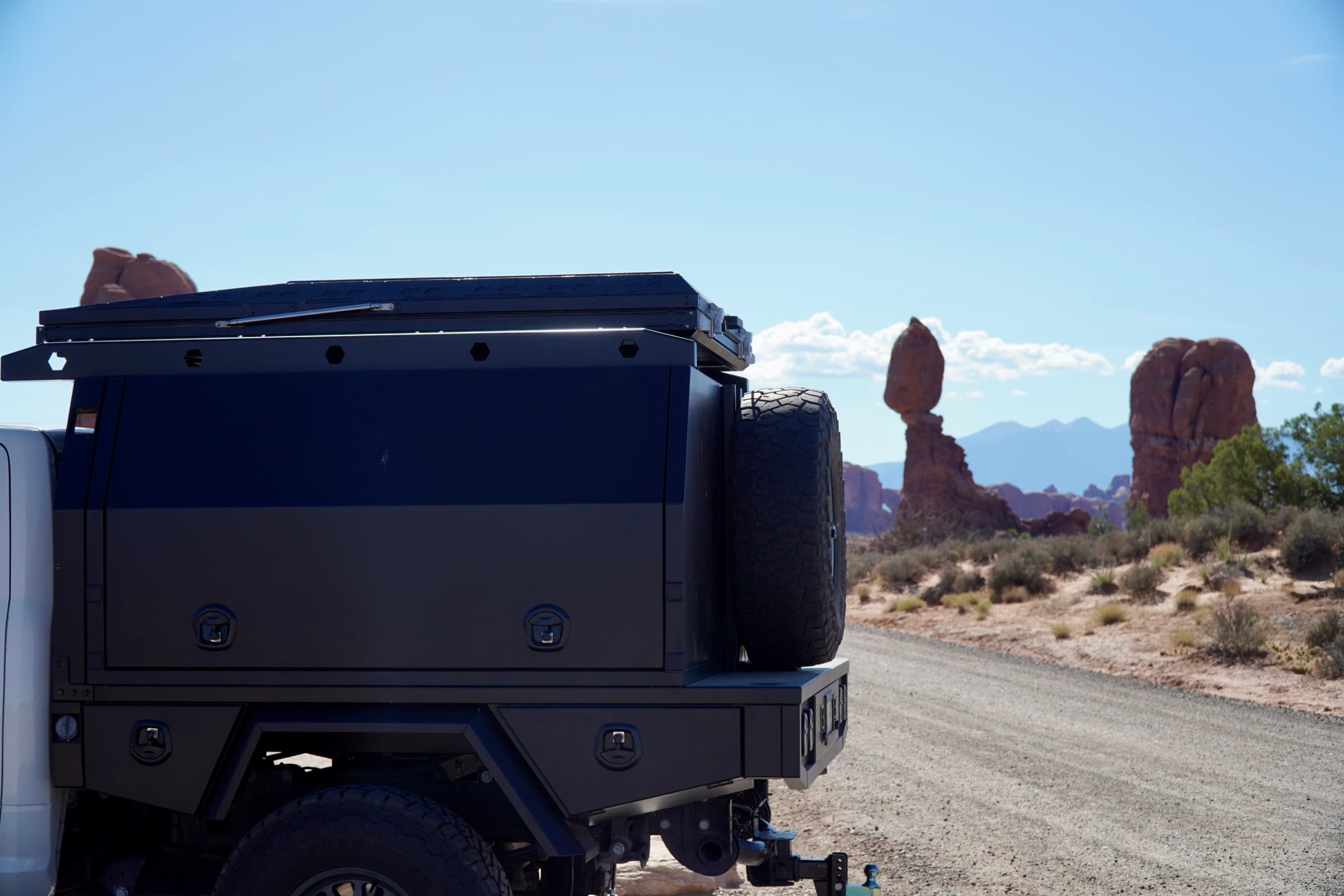 Ram 3500 with a black aluminum touring canopy and rooftop tent parked on a gravel road at Arches National Park, Balanced Rock in the background