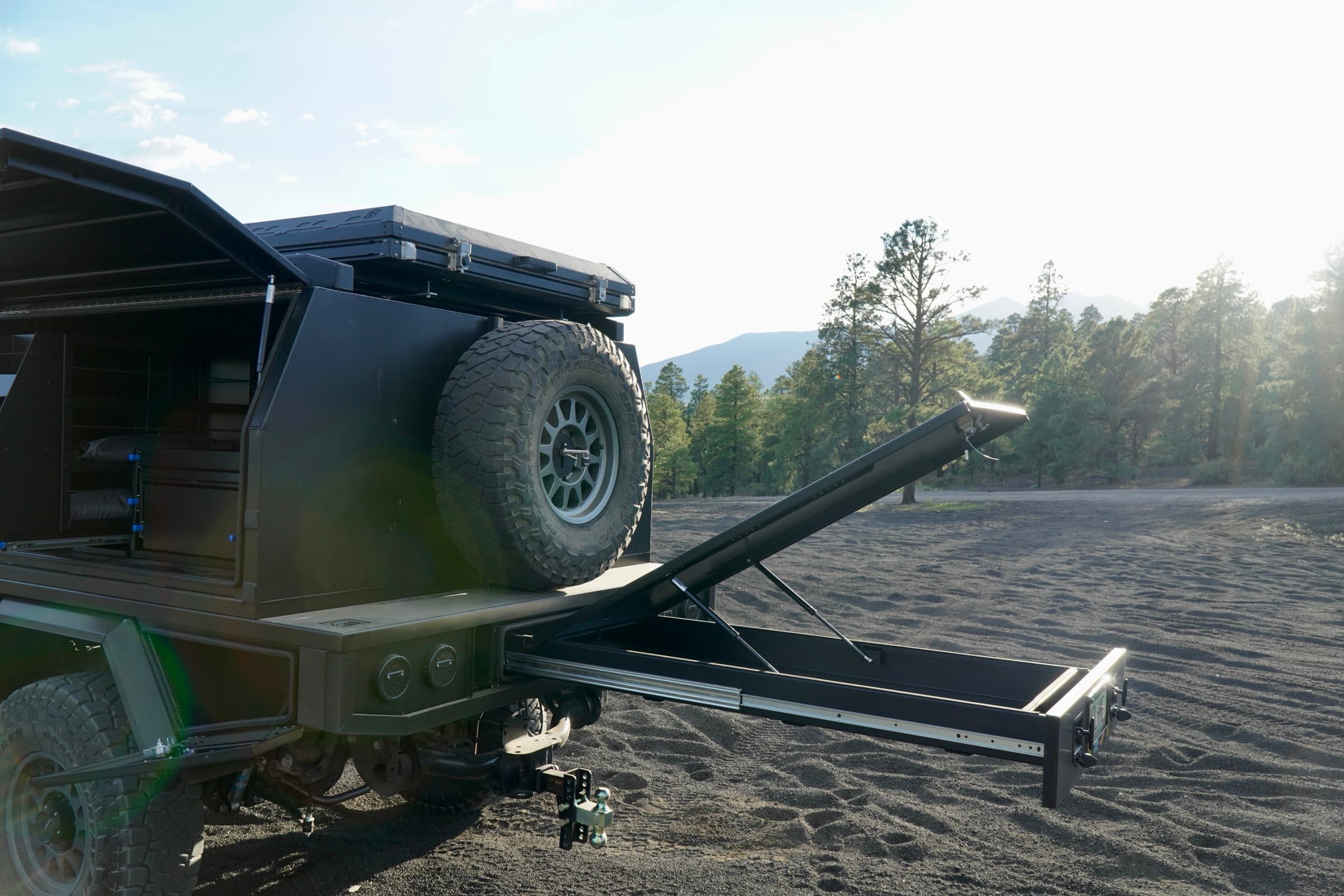 Rear of the Ram 3500 touring canopy build with the full-width trundle drawer fully extended and its gas-strut lid raised, parked in a pine forest
